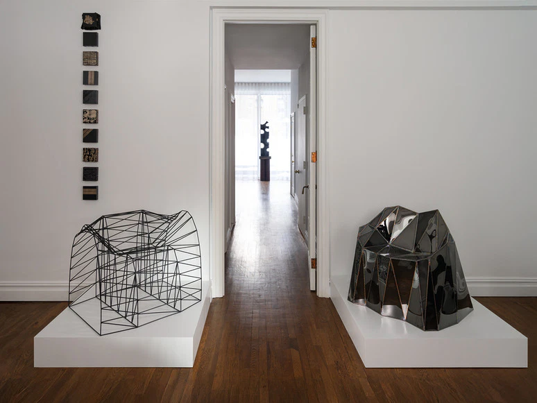 art gallery room displaying abstract sculptures on white platforms, framed by a bright doorway leading to another part of the gallery