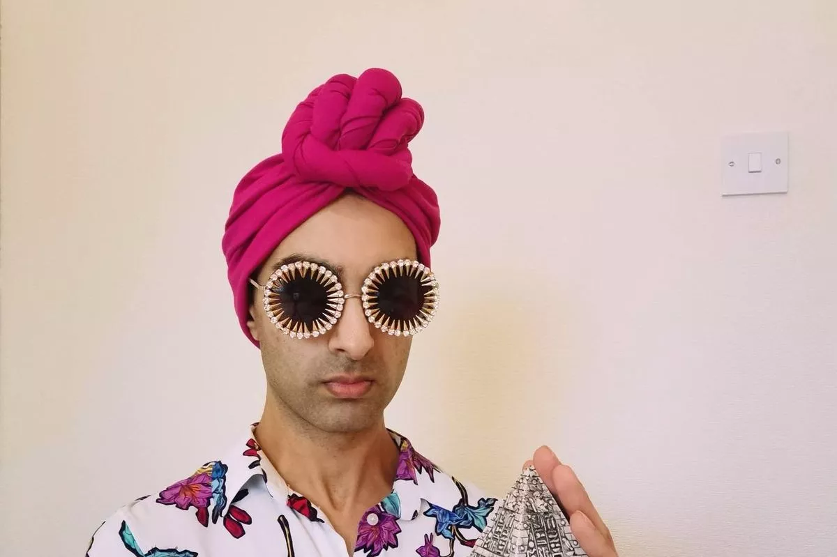 An individual dressed in a floral shirt and large round sunglasses, wearing a pink headscarf, is holding a small, metallic object. The background is plain white.