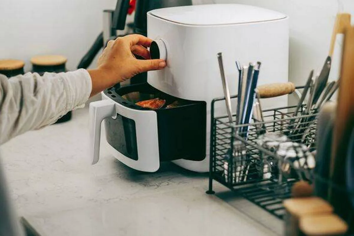 Person adjusting air fryer with visible drawer containing food in modern kitchen, organized countertop with utensils nearby.