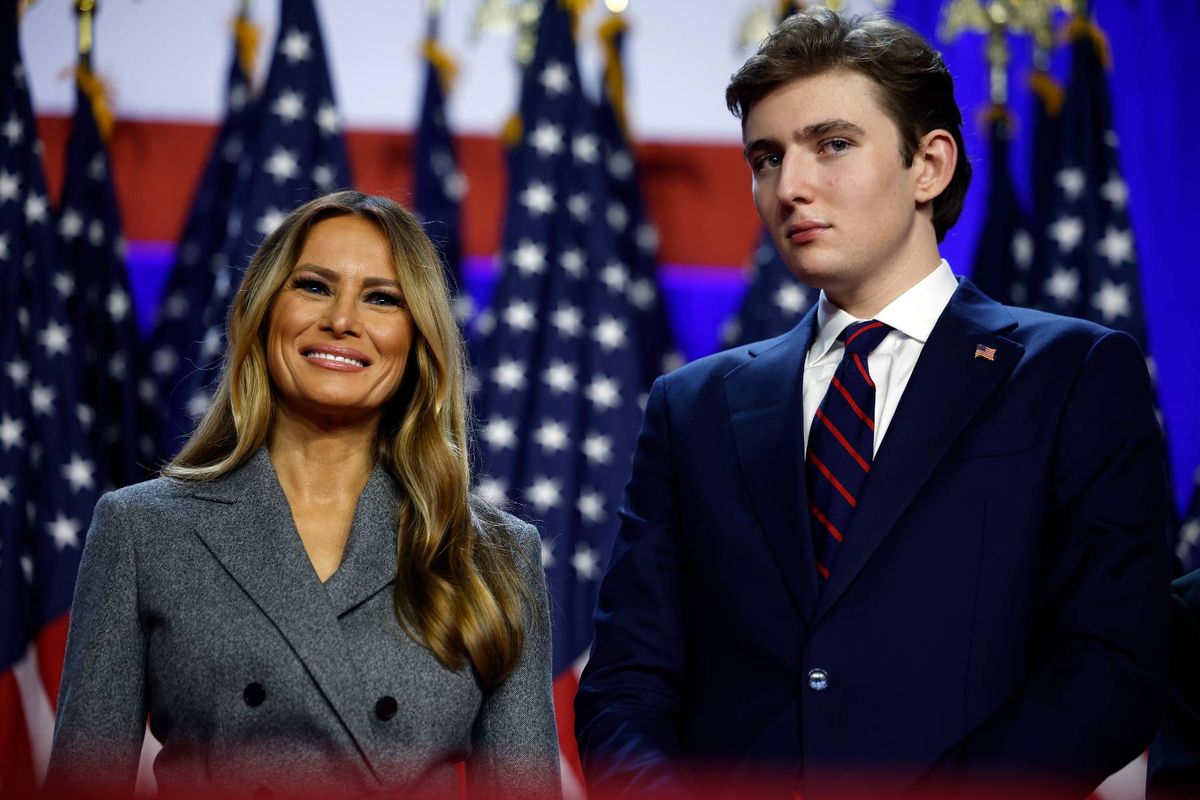 A formal event is taking place with a backdrop of American flags. A woman and a man, both dressed in formal attire, are standing together, possibly addressing an audience.