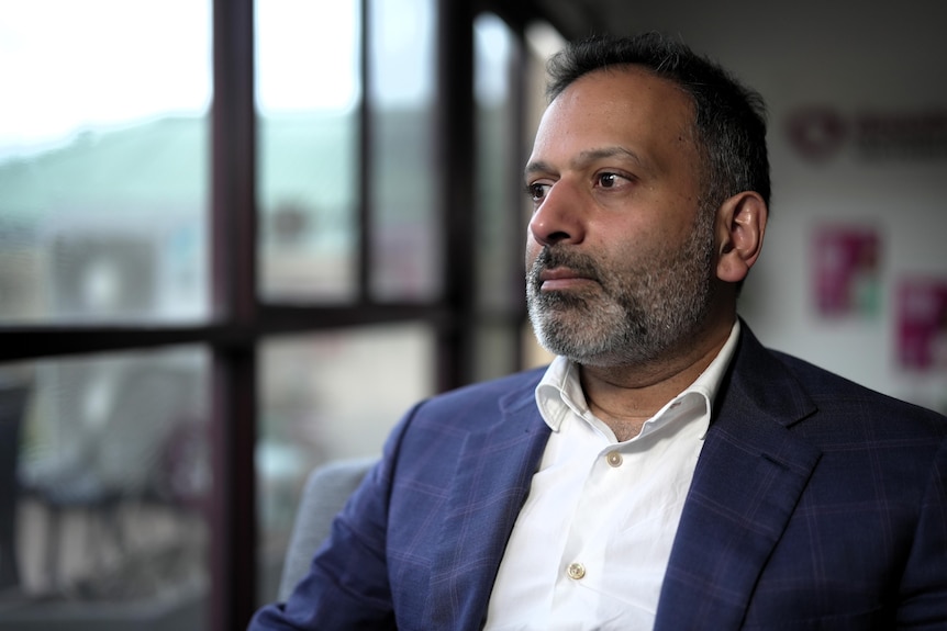 A man of south Asian background with short brown hair and a short beard sitting in an office