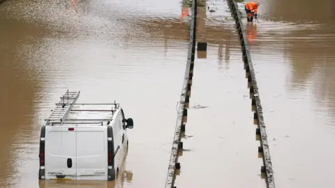 PA Media A white van is stranded in floodwater on the A189 Spine Road near Blyth, Northumberland. The water comes up to the the top of the vehicle's number plate. A worker in an orange high-vis uniform is crouched down further up the road.