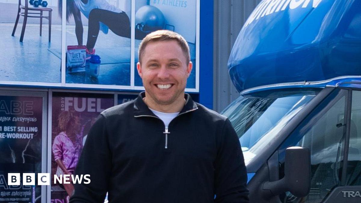 Tom Ryder, who is smiling at the camera, is standing in front of van with Applied Nutrition branding and holding an Applied Nutrition branded water bottle.