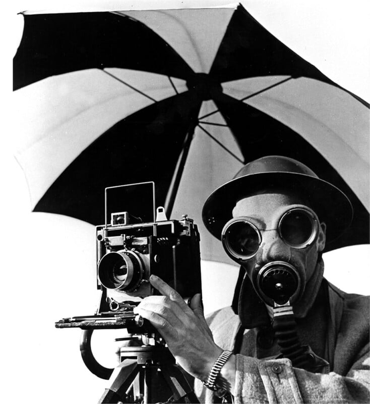 A person wearing a hat, large goggles, and a gas mask operates an old-fashioned camera under a large, open black-and-white umbrella.