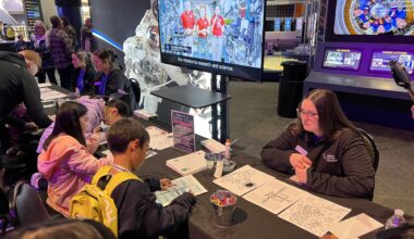 Children participate in a hands-on space activity at Space Center Houston, placing stickers on a poster at an interactive table while visitor center team members provide guidance.