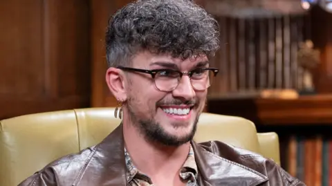 BBC/Studio Lambert/Euan Cherry Jack from Traitors is smiling while sat in a library and wearing a brown leather jacket