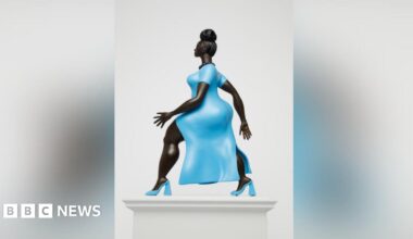 A statue of a woman of colour wearing a striking baby blue dress with matching high heels of the same colour. She has her hair up in a bun and is wearing a black beaded necklace. She is walking with purpose with one arm in front of the other.