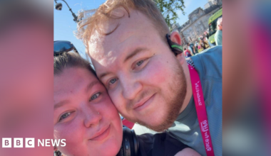 A close up selfie of Richard with his girlfriend, Sophie at the Race for Life running event. Sophie, on the left, has brown hair and wears sunglasses on the top of her head. Richard is standing next to Sophie with his cheek touching her cheek. He wears a green t-shirt, a pink medal, earphones and has short blonde hair.