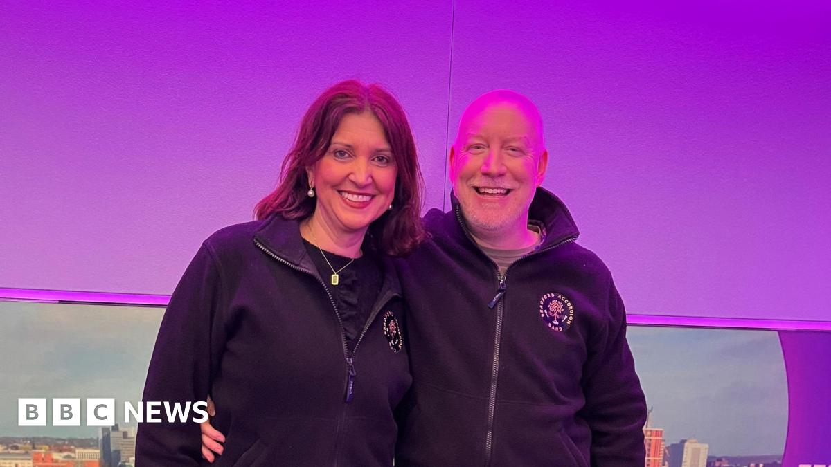 A woman with dark hair hugging a bald man man in a radio studio with a purple backdrop