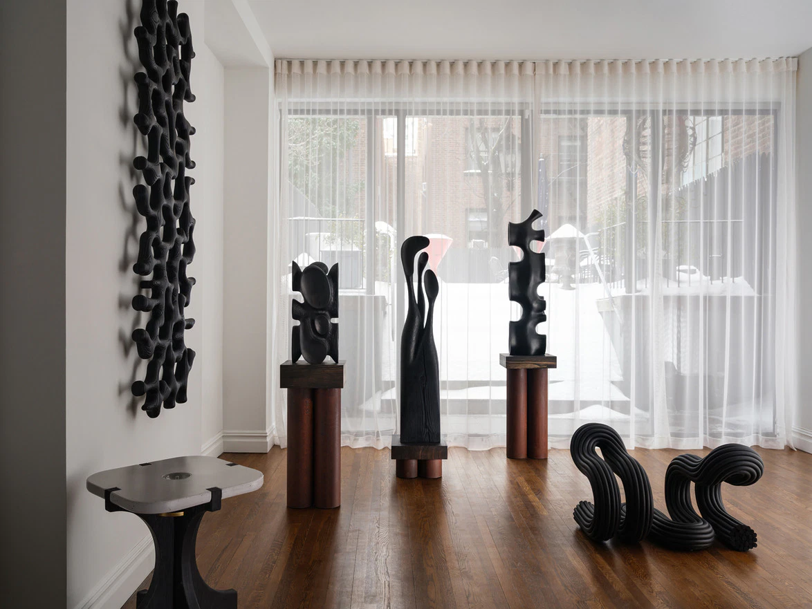 Contemporary room with abstract black sculptures on wooden stands and a textured wall piece, with large windows in the background.