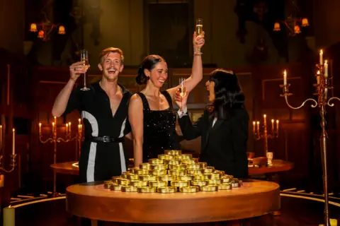 Stephen, Rachel and Claudia Winkleman holding up glasses of champagne and smiling behind a table with a pile of gold coins