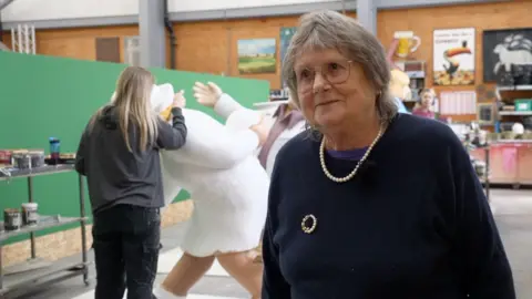 A woman with grey hair and glasses smiles at the camera. She is stood in front of a sculpture being painted behind her.