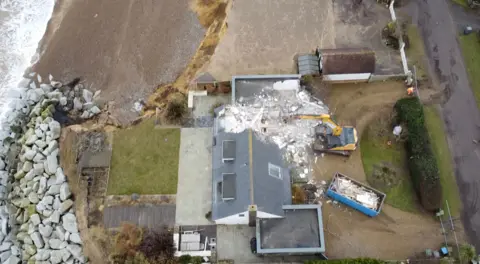 Jamie Niblock/BBC A drone image above a property on the coast that is being demolished. White rubble from the home can be seen around it. 