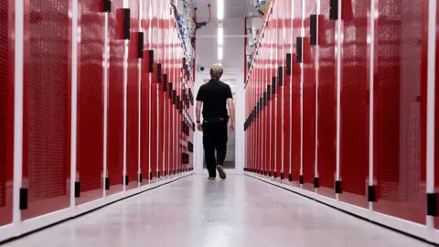 AFP via Getty Images A worker at a data centre in Sydney, Australia, walking down the middle of two rows of red-faced computer servers