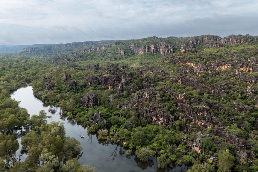 An aerial shot of sandstone country, where a small boat can be seen travelling along a river snaking past green trees.