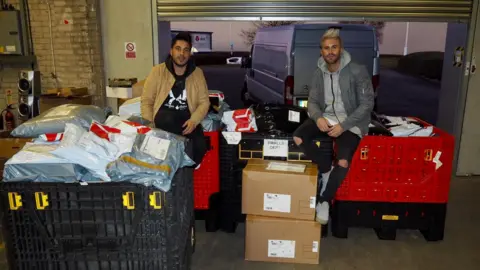 The Couture Club Ross Worswick and Scott Shashua are sitting on top of large crates filled with parcels inside a small warehouse.