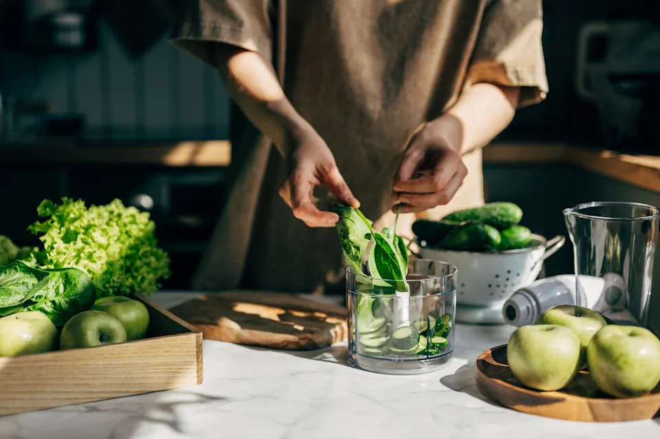 Person preparing a green smoothie, adding spinach and cucumber to a blender. Fresh apples and lettuce are on the counter