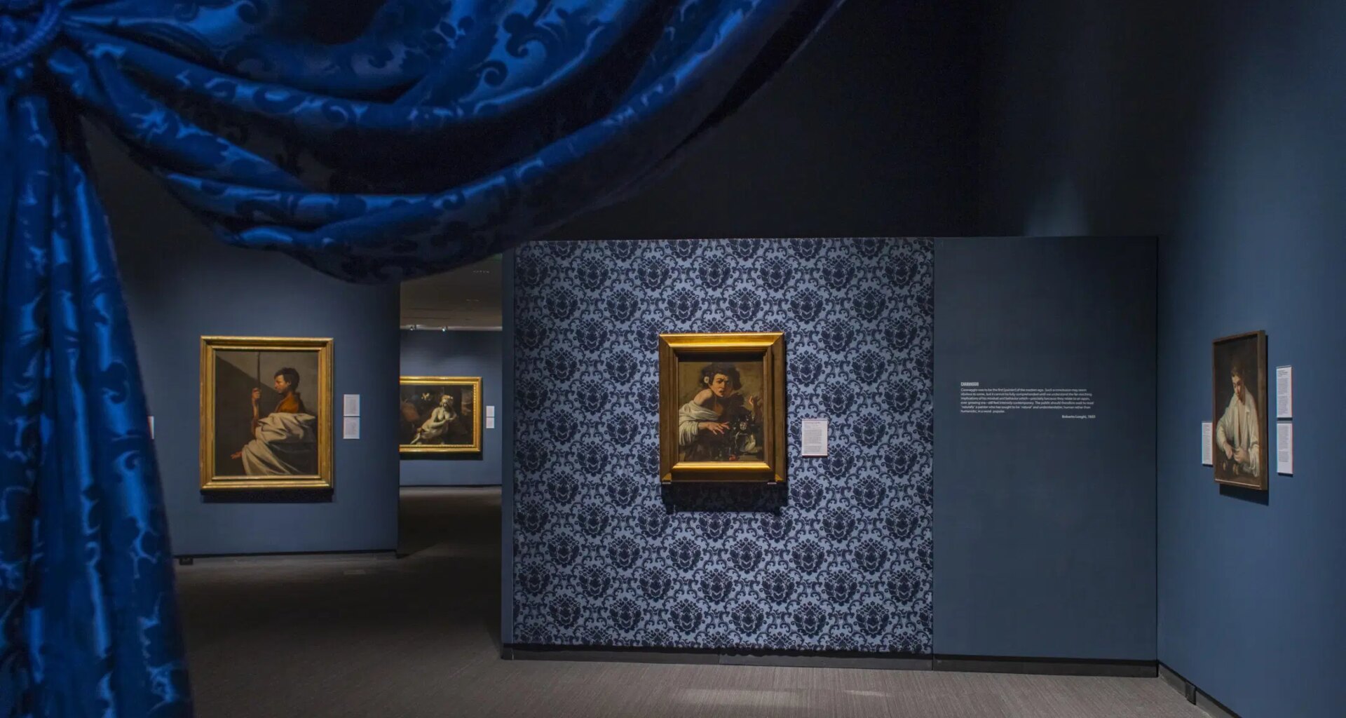 Interior view of an art gallery with framed paintings on blue patterned walls and a blue curtain in the foreground.
