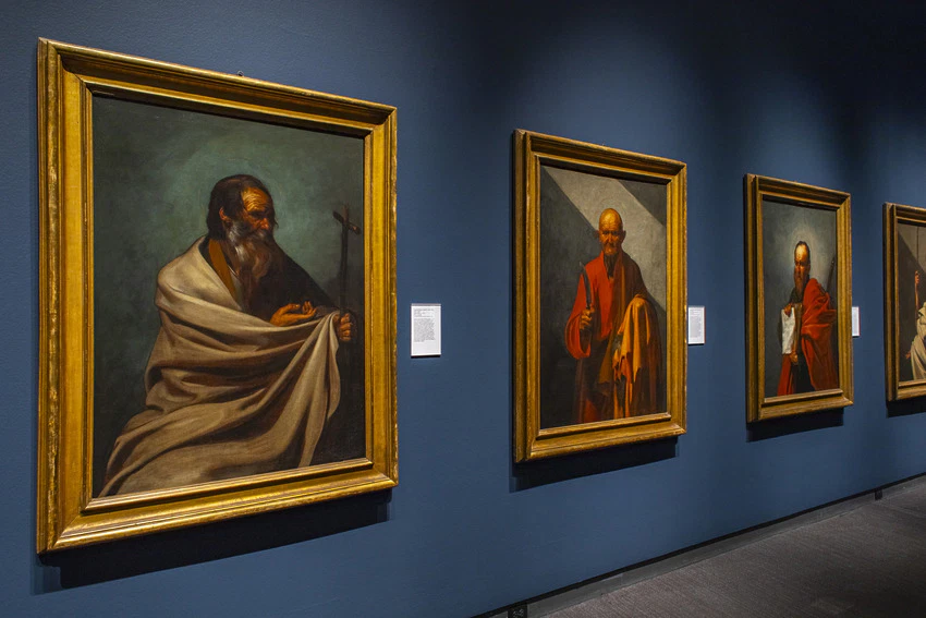 Art gallery with framed paintings of historical figures on a blue wall, showcasing detailed portraits in a dimly lit setting.