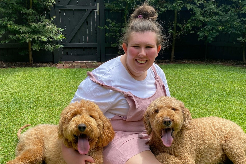 A young woman sits on the grass with two dogs