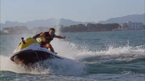 FCA/Pensions Regulator A young man in sunglasses races across the sea on a jet ski in front of a holiday destination coastline.