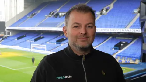 Jamie Niblock/BBC Paul Hamilton standing in a box at Ipswich Town FC ground. He has short greying hair and short greying beard and is wearing a black zipped up top with HaloITSM written on the left side. Beyond him are rows of blue seats and the football pitch.