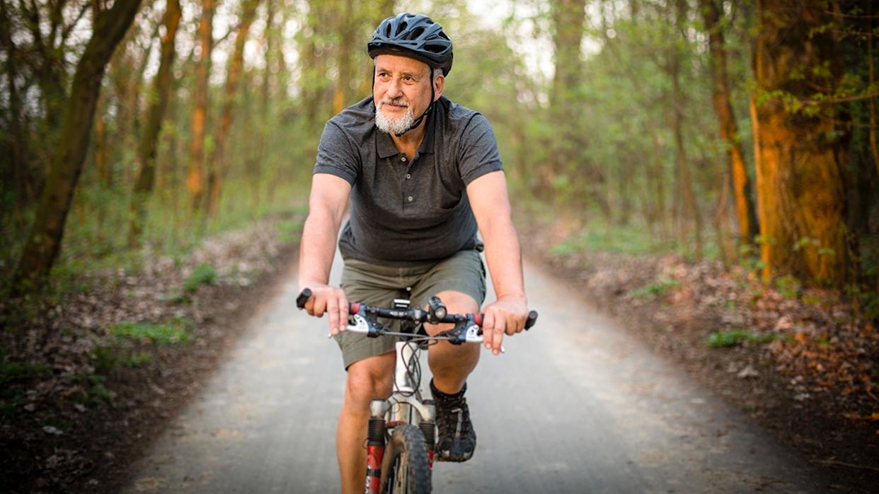 An elderly man rides a bicycle in the woods while wearing a helmet.