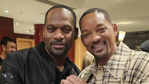 Shad Ellis Will Smith standing next to a man who looks very similar facially. Both are smiling. The lookalike is wearing black with the letters "MIB" on his right arm. Will Smith is wearing a light check pattern outfit.
