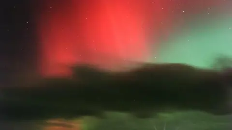 BBC Weather Watchers/Hol Red and green aurora colours appear in the night sky above a dark landscape.