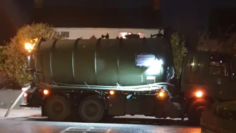 Nita Brown A tanker in operation at night pumping sewage out of a manhole in the road. There are lights on the tanker, and a house in the background has a light on upstairs.