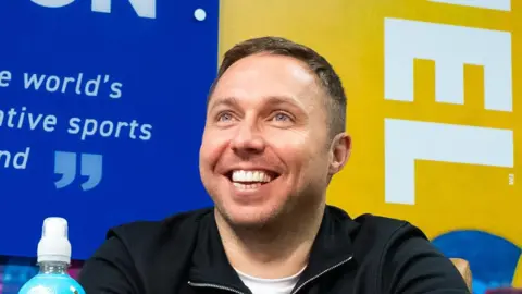 Applied Nutrition Tom Ryder, who is smiling, is sitting a desk in front of a range of Applied Nutrition products. There are a number of Applied Nutrition advertisements in the background. 