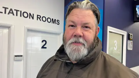 Nicola Haseler/BBC A man with a grey beard and hair looks at the camera. He wears an olive green rain jacket and is standing in front of a sign that reads "CONSULTATION ROOMS" in a pharmacy. 