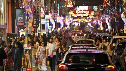 AFP via Getty Images Shoppers walk along Commercial Street in Bengaluru city, illuminated with decorative lights ahead of New Year celebrations on 30 December, 2025. 

