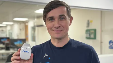 Owlstone co-founder Billy Boyle is wearing a navy blue t-shirt and holding up a diagnostic breath test for the camera. It is pale grey and the size and shape of a computer mouse. Boyle is smiling. He has dark hair and is standing alongside a window which gives a view into the company's diagnostic labs. 