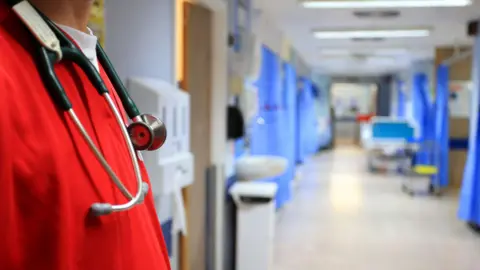 PA Media A stock image of a hospital ward. The image shows an empty ward with several bays with blue curtains, which are blurred in the background. In the forefront of the image is a man in bright red scrubs with a stethoscope around his neck. 
