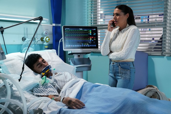 Priya, on the phone, stands over an unconscious Nugget in hospital in EastEnders