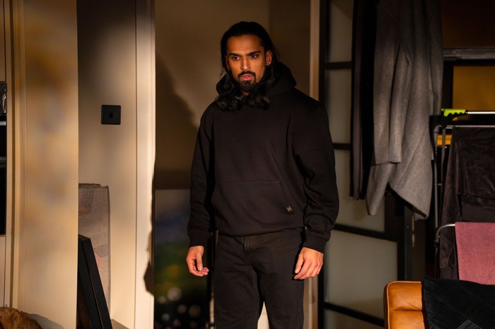 543576,TITLE:Eastenders Ravi Gulati, with his hair down and wearing a black hoody, stands at the bottom of a staircase in EastEnders