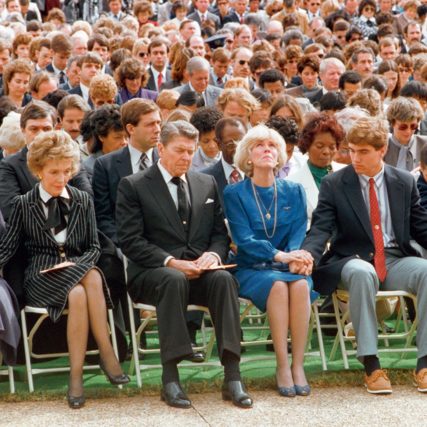 Colour photo of the front row of a large seated crowd showing well dressed mourners mostly looking down or skyward.