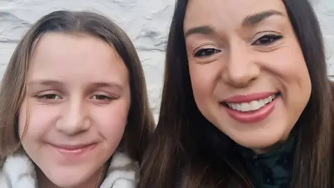Family photo Eliza, aged 11 and her mother Sorraya. Both are white with long light brown hair and are smiling at the camera. Both are wearing makeup. There is a white outdoor stone wall behind them. The collars of coats are just visible.