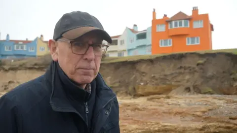 Jamie Niblock/BBC Richard Bennett stands at the bottom of an eroded cliff where a row of colourful homes rest at the top. He wears cap with a navy coat and fleece and glasses.