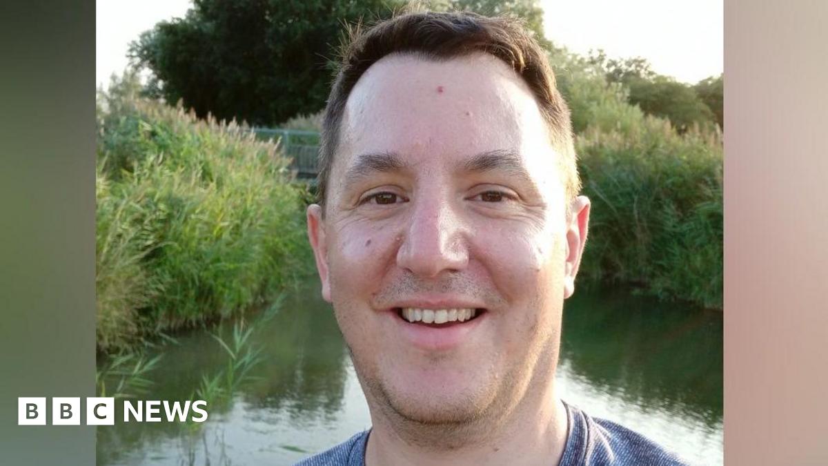 James Clements is standing in front of a small lake which is surrounded by tall grass. He has short brown hair, and is smiling at the camera. He is wearing a blue T-shirt.