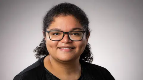 Dr Nina Higson-Sweeney A professional headshot of Dr Nina Higson-Sweeney wearing a black t shirt and black rectanglar framed glasses 