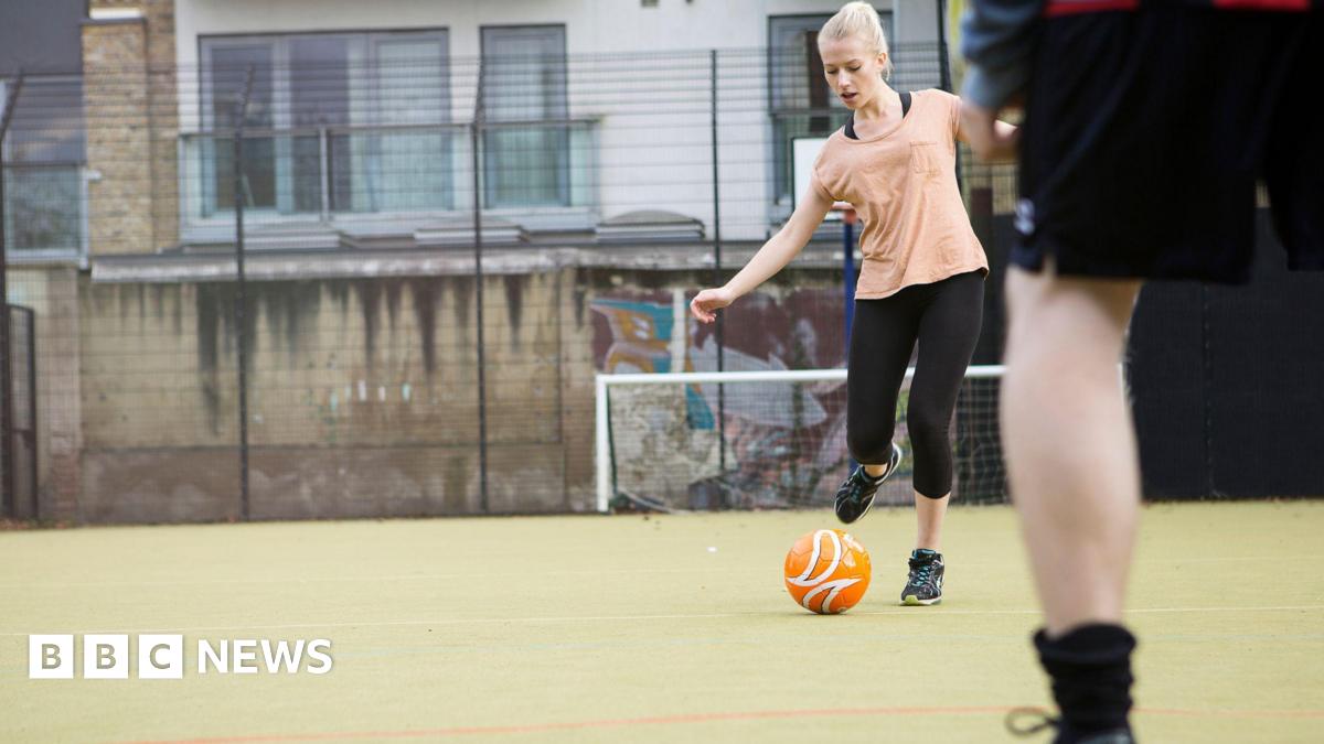 A woman with white blonde hair in a ponytail and a peach T-shirt with black leggings is kicking a ball on an outdoor football pitch. Someone blurred in the foreground is standing in front of her.