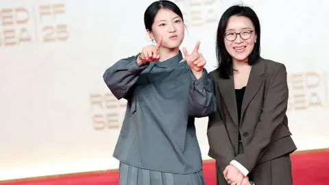 Getty Images Seo Su-bin and Yoon Ga-eun attend The World of Love screening. Seo, on the left, points both index fingers at the camera, while Yoon, standing next to her, fold her hands in front of her body and smiles at the caemra.
