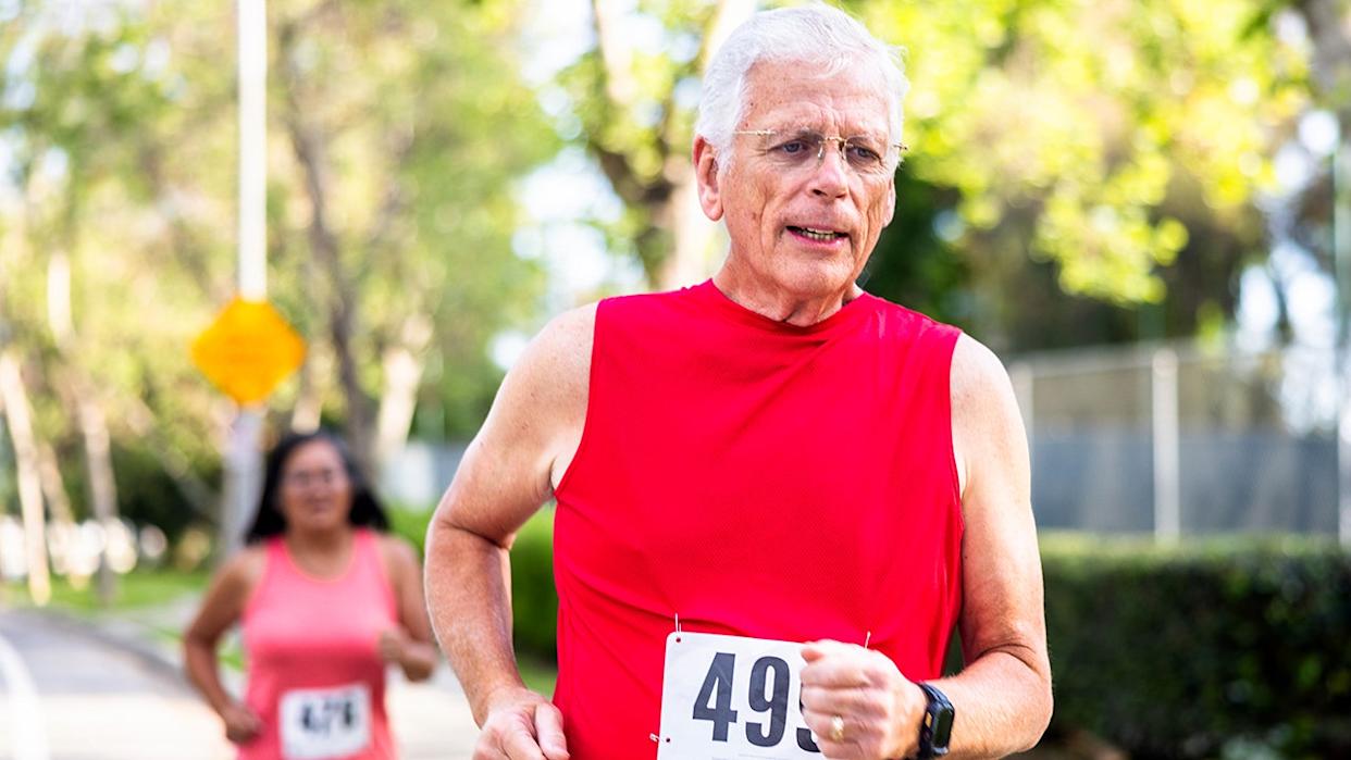 An elderly man with white hair and wearing a red shirt and glasses runs in a race.