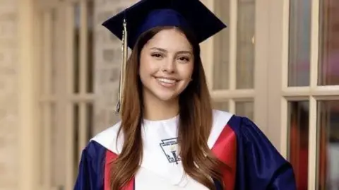 Bexar County Sheriff's Office Camila Mendoza Olmos, with long brown hair, smiles and poses for a photo in a red, white and blue graduation cap and gown