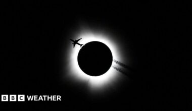 Black sky with white halo around a black disc. Silhouetted plane travelling through white halo