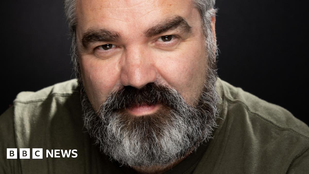 A man with a black and grey beard wearing a khaki green shirt