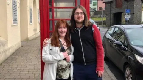 Lewis Relfe Ameila and Lewis Relfe stood in front of a red phonebook. She is wearing a grey hoodie and a black top with a pattern on it. He is wearing a black and red hoodie.