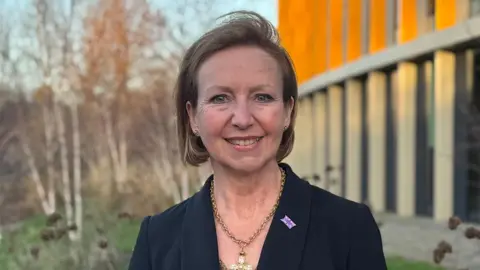 Jane Hutchins is standing outside the Bradfield Centre at the heart of the science park. The bright orange external pillars of the building are to the right of the photo, with trees and greenery to the left. Jane is in the middle, smiling at the camera. She has dark blonde hair in a chin length bob, being blown slightly by the wind. She is wearing a black suit jacket and a chunky golden necklace with a sparkly pendant.